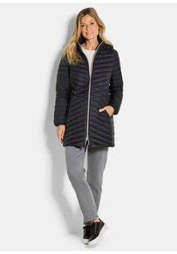 Zwarte gewatteerde jas met een hoge kraag, ritsluiting en zijzakken. Draagt met grijze broek en zwarte sneakers. Soepele textuur.