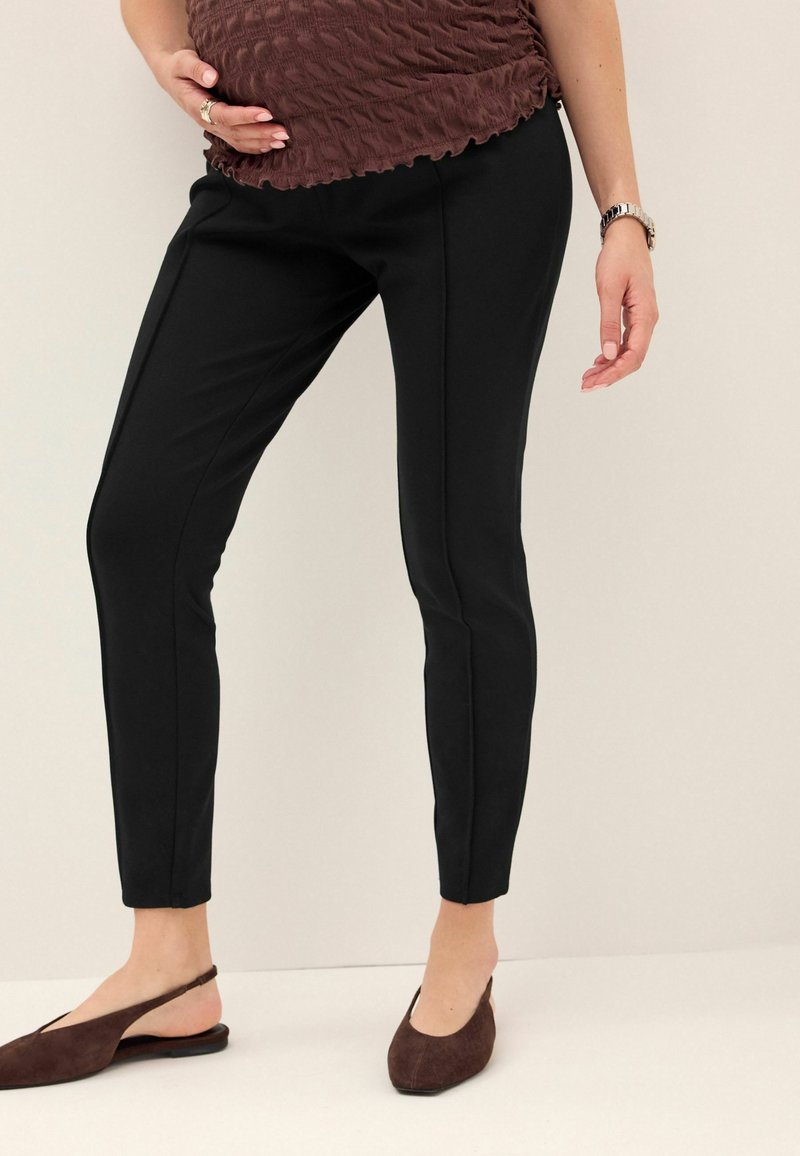Pantaloni neri aderenti con una texture liscia, caratterizzati da cuciture laterali. Indossati con un top marrone strutturato e scarpe slip-on marroni.