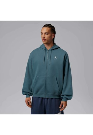 Mann mit geflochtenem Haar, der einen türkisfarbenen Hoodie mit Reißverschluss, Kapuze und vorderen Taschen trägt, steht vor einem einfarbigen grauen Hintergrund.