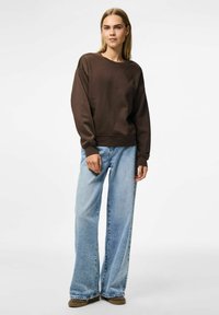 Sudadera marrón de corte relajado, con puños anchos y cuello redondo, combinada con jeans de pierna ancha de color azul claro y calzado marrón.