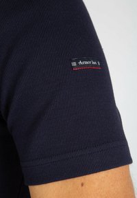 T-shirt in tessuto a righe di colore blu navy con maniche corte. Presenta un'etichetta nera con accenti rossi sulla manica sinistra alta.