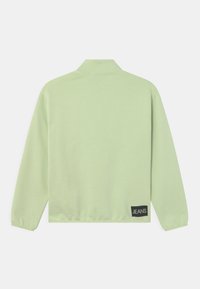 Pullover verde chiaro con colletto polo, maniche lunghe e polsini elasticizzati. Presenta un'etichetta nera "JEANS" nella parte bassa a sinistra. Tessuto morbido.