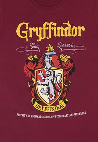 Bordeauxrotes T-Shirt mit einem Gryffindor-Wappen, das einen Löwen, einen Ritterhelm und gelben Text zeigt, entworfen für Quidditch-Fans von Hogwarts.