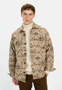 Tan gestreepte shirtjack met een geometrisch ontwerp in bruintinten, voorzien van twee borstzakken en knoopsluitingen. Gelaagd over een crèmekleurige coltrui.