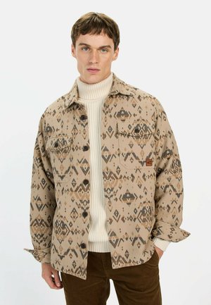 Tan gestreepte shirtjack met een geometrisch ontwerp in bruintinten, voorzien van twee borstzakken en knoopsluitingen. Gelaagd over een crèmekleurige coltrui.