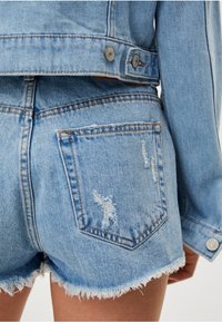 Veste en denim bleu clair avec un ourlet brut et un short déchiré, dotée d'une fermeture à boutons et de coutures visibles sur les poches.