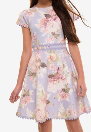 Mädchen trägt ein lavendelfarbenes Kleid mit rosa-weißem Blumenmuster, gezacktem Abschluss an Ärmeln, Taille und Saum, steht mit entspannten Armen.