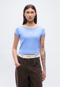 Ανοιχτό μπλε, εφαρμοστό βαμβακερό t-shirt με κοντά μανίκια και στρογγυλή λαιμόκοψη. Διαθέτει μικρό λογότυπο Calvin Klein. Συνδυάζεται με σκούρο καφέ παντελόνι.