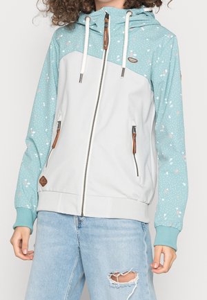 Person iført en hvid og lyseblå plettet lynlåshættetrøje med snore og lyseblå revnede jeans.