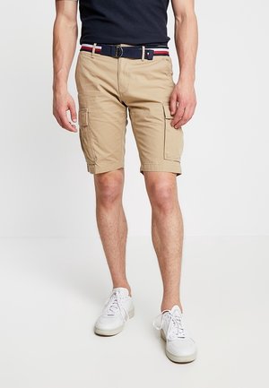 Pantalones cortos de cargo en color caqui con múltiples bolsillos, combinados con un cinturón azul marino con rayas rojas y blancas. Llevados con zapatillas blancas.