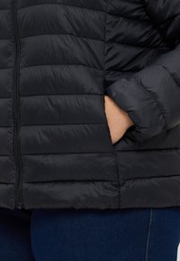 Melns piepūšamais jaka ar iepakojumu, priekšējo rāvējslēdzēju un sānu kabatām; izgatavota no viegla, mirdzoša materiāla; piedurkņu gali ar ribota auduma apdari.