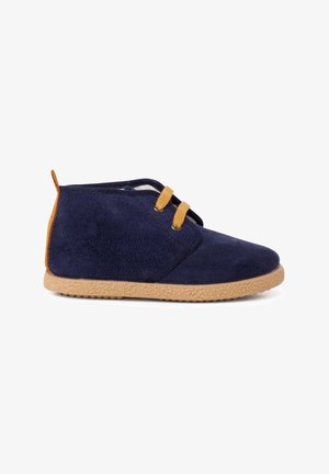 Botă din suede bleu marin cu o talpă din cauciuc bej deschis. Prezintă două șireturi galbene și un tab cu buclă la spate pentru o purtare ușoară. Design cu tăietură joasă.