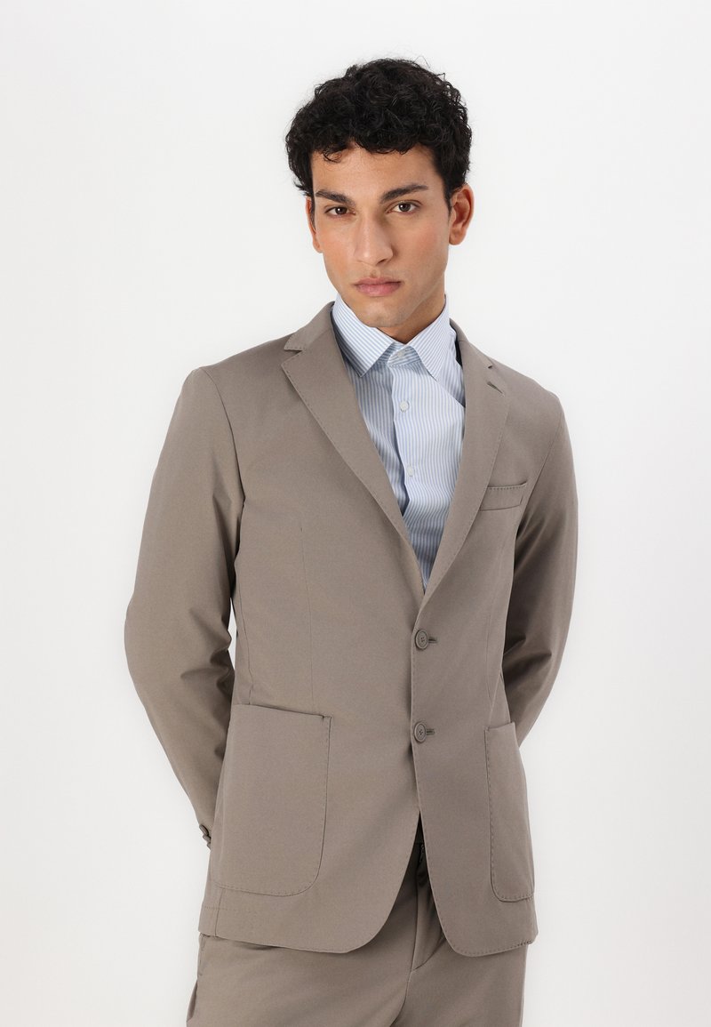 Veste de costume beige en tissu lisse, avec deux poches avant, un revers cranté et deux boutons, associée à une chemise à rayures bleu clair.