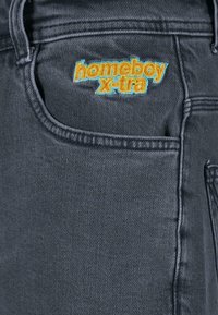 Jean en denim noir avec broderie "homeboy x-tra" en bleu vif et orange sur la poche avant, ornés de détails de couture standard.