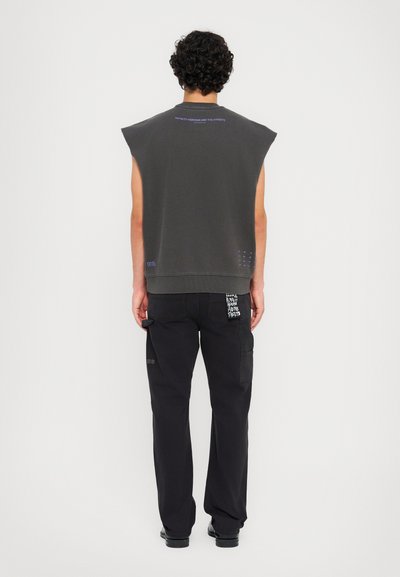 T-shirt sans manches gris avec un col côtelé et un texte violet, associé à un pantalon cargo noir avec des détails graphiques blancs et des poches.