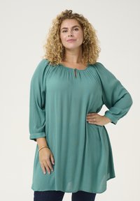 Blusa turchese con scollatura a chiave e dettagli plissettati. Presenta maniche a tre quarti e una vestibilità rilassata, realizzata in un tessuto morbido e leggero.