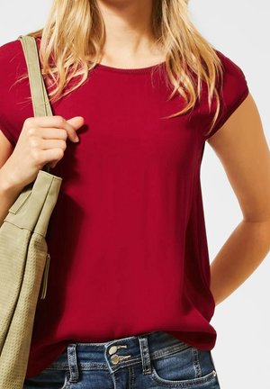 Frau mit langen blonden Haaren trägt ein rotes Kurzarmshirt und blaue Jeans, sie hält mit der linken Hand den Riemen einer beigen Schultertasche.