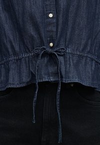 Σκούρο μπλε πουκάμισο από τζιν με λευκά κουμπιά, δεμένο με φιόγκο στη μέση, σε συνδυασμό με μαύρο παντελόνι που δείχνει τις εμπρόσθιες τσέπες και τη μέση.