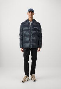 Dunkelblauer Steppjacke mit Reißverschluss vorne, breiten gesteppten Abschnitten, Seitentaschen und einem hohen Kragen. Getragen mit schwarzen Jeans und beige Sneakers.