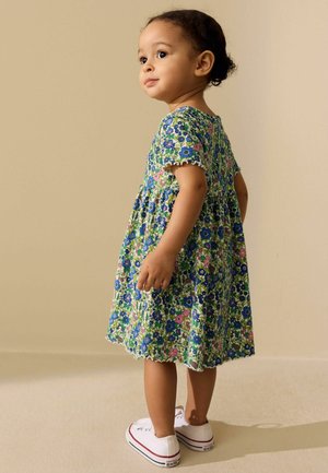 Småbarn med mørkt krøllet hår iført en blå, grøn og pink blomsterkjole og hvide sneakers, stående og kigger til siden.