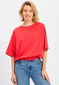 Rode top met korte mouwen en ronde hals, in een ontspannen fit, gecombineerd met blauwe denim jeans. Het materiaal lijkt zacht met een gladde textuur.