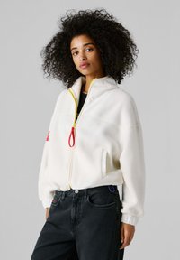 Λευκό fleece μπουφάν με ψηλό γιακά, κίτρινο φερμουάρ και κόκκινο τράβηγμα. Διαθέτει πλαϊνές τσέπες και ριμπ μανσέτες· συνδυάζεται με μαύρο τζιν.