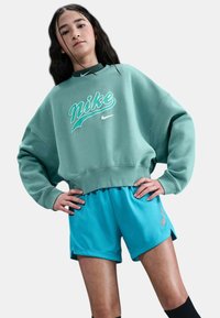 Giovane che sta in piedi con le mani sui fianchi, indossando una felpa Nike di colore teal e pantaloni Nike blu brillante, su uno sfondo neutro.