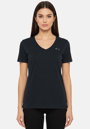 Donna in piedi rivolta in avanti che indossa una maglietta scura semplice con scollo a V e maniche corte, con un piccolo logo sul petto a sinistra, su sfondo bianco.