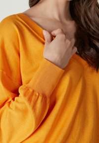 Ljusorange stickad tröja med V-ringning och samlade ärmar. Tyget är mjukt med en lätt strukturerad yta. Bekväm och avslappnad design.