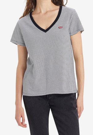 Kvinde iført en hvid og sort stribet V-hals T-shirt med et lille rødt logo på brystet, kombineret med sorte jeans, foran en hvid baggrund.