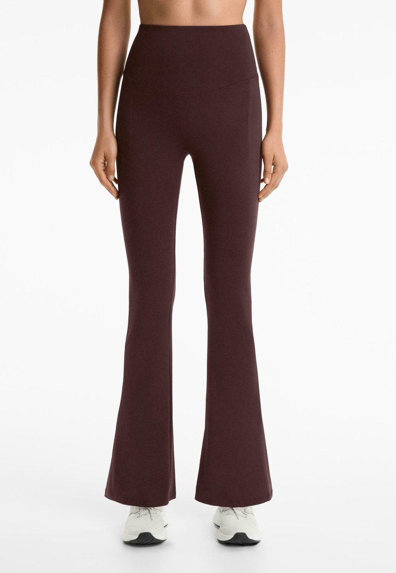 Leggings flare taille haute bordeaux avec une texture lisse et des détails de couture, associés à des chaussures de sport blanches.