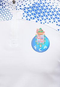 Λευκή αθλητική φανέλα με μπλε γεωμετρικό τρίγωνο κοντά στον γιακά και μπλε κυκλικό έμβλημα FCP στο στήθος.