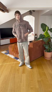 Ανοιχτό καφέ φούτερ με κείμενο "LUCID DREAMS", ανοιχτό τζιν και ασημί παπούτσια. Στεκόμενος σε ένα σαλόνι με ένα φυτό και πορτοκαλί καναπέ.