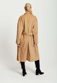 Long, tan wool coat with a belted waist, wide sleeves, and a back vent. Worn over black pants and ankle boots. Smooth texture.