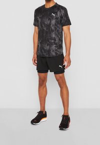 Mies, който носи черен спортен екип на Puma, включващ тениска с модел на дим, наслойка от шорти и черни маратонки с оранжеви акценти.