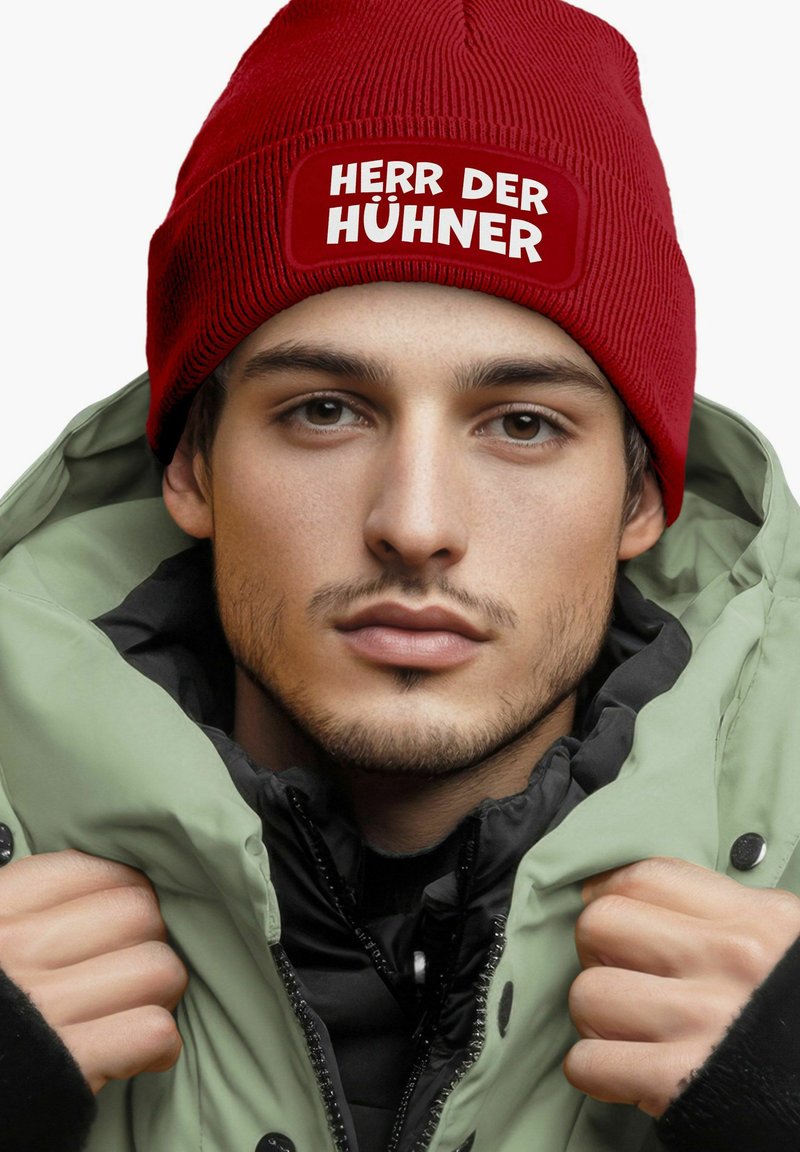 Red ribbed beanie with white text "HERR DER HÜHNER," paired with a black jacket and a light green outer coat, featuring a high collar.