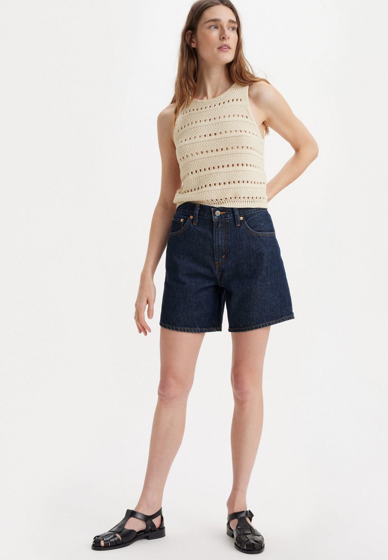 Un débardeur en tricot beige clair avec des motifs ajourés, associé à un short en denim bleu foncé à cinq poches et à l'ourlet ajusté. Sandales noires.