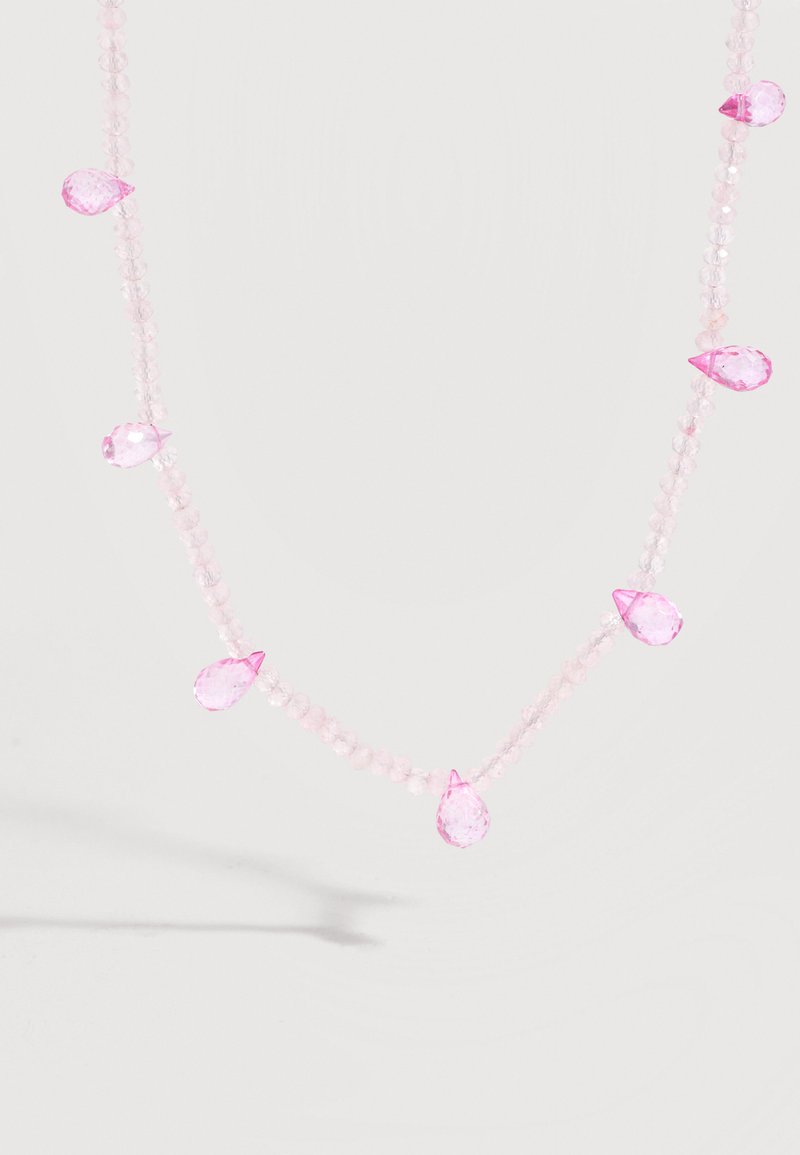Collier délicat avec des perles roses translucides et de plus grosses pierres précieuses roses en forme de goutte espacées le long du fil sur fond blanc.