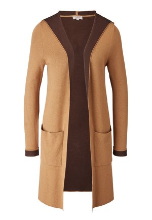 Vrouw draagt een lange cardigan in beige en donkerbruin, een witte coltrui, crèmekleurige joggingsbroek en witte hoge sneakers.