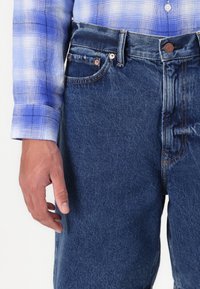Denim jeans in dark blue with a fringed edge, featuring copper hardware and contrasting orange stitching. Sleeves of a blue checkered shirt visible.