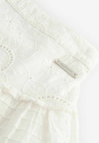 Vit bomullstyg med broderade blommönster, spetskant och en silverfärgad metalltagg med texten "ALLSAINTS." Texturerade volanger i botten.