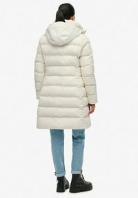 Roomkleurige donsjas met horizontale stiksels, capuchon en een middenlange snit. Draagt over lichtblauwe jeans en zwarte veterlaarzen.