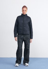 Veste matelassée noire avec un col haut, associée à un pantalon noir ample orné de motifs floraux. Le mannequin se tient devant un fond blanc.