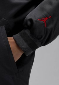 Czarna sportowa marynarka z czerwonym haftowanym logo na rękawie, prążkowanymi mankietami i gładką fakturą oraz kieszenią na zamek.
