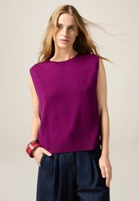 Fuksjowy bezrękawnik z miękkiego dzianego materiału, z okrągłym dekoltem i bocznymi rozcięciami, zestawiony z luźnymi ciemnymi spodniami dżinsowymi.