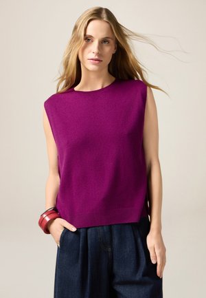 Fuksjowy bezrękawnik z miękkiego dzianego materiału, z okrągłym dekoltem i bocznymi rozcięciami, zestawiony z luźnymi ciemnymi spodniami dżinsowymi.