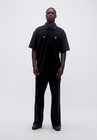 Άνδρας όρθιος, φοράει μαύρο φαρδύ πόλο μπλουζάκι και μαύρο παντελόνι, μπροστά σε απλό λευκό φόντο.