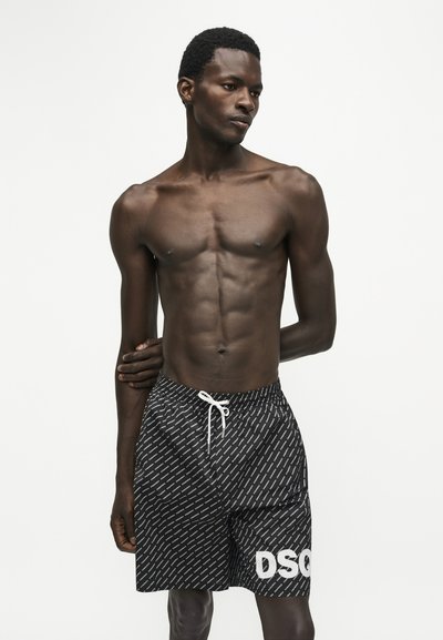 Homme torse nu portant un short à motifs noir et blanc avec un cordon blanc, debout, une main dans le dos, sur fond blanc.