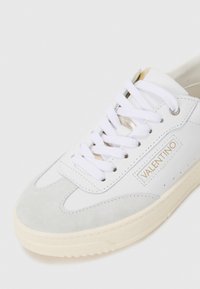 Sneakers bianchi in pelle con dettagli in suede grigio, lacci bianchi, un logo dorato e una suola in gomma texturizzata color crema.