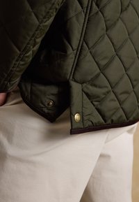Gesteppte dunkelgrüne Jacke mit messingfarbenen Druckknöpfen, getragen über hellbeigen Hosen, zeigt strukturierte Nähte und Details am Jackensaum.
